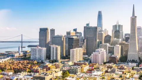 Getty Images A panoramic view from above San Francisco's financial district on a bright sunny day. Tall buildings loom over a lower cityscape, and the famous Golden Gate Bridge is seen in the background.