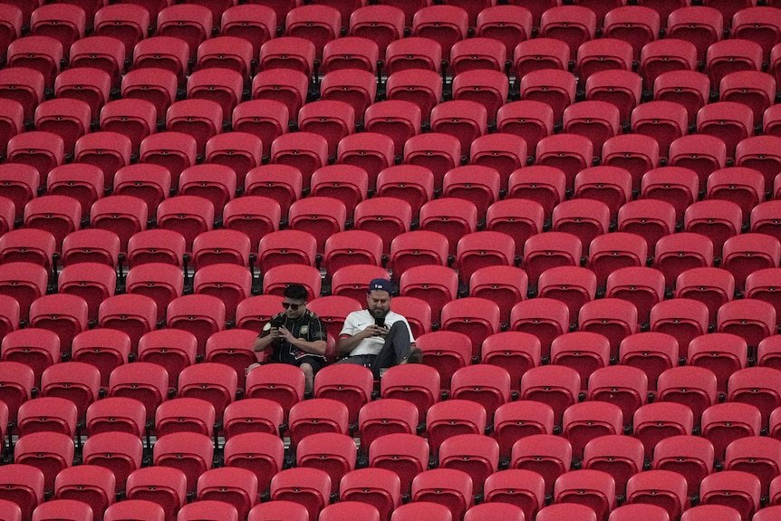 Two men sitting amid dozens of empty rows of red stadium seating, with one looking at a phone with a leg up