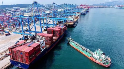 Getty Images Aerial view of a container terminal in Qingdao with blue cranes hovering over container ships and a smaller green and orange boat approaching in east China's Shandong province Monday, 11 August, 2025.