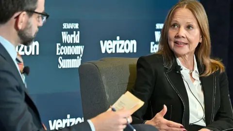 Getty Images Mary Barra in white shirt and black suit jacket with white trim gestures while speaking to a man in glasses in front of a background reading "World Economy Summit".