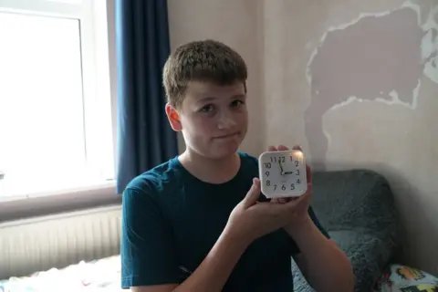 A teenage boy stands holding up an analogue alarm clock.