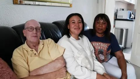A dad and mum sit on a sofa, with their teenage daughter sat between them. They are all smiling at the camera.