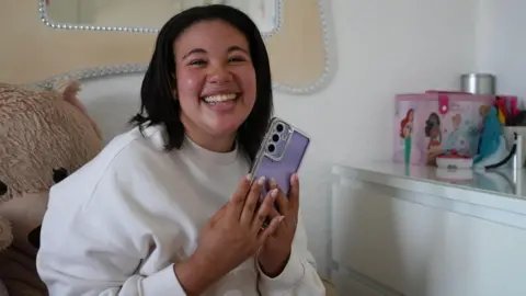 BBC A teenage girl clutches her phone which has a purple phone case and smiles while sitting on her bed in her bedroom.