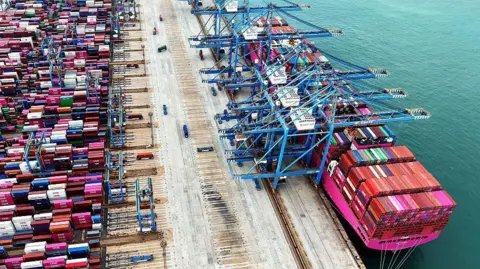 Getty Images Overhead view of a container ship in a container port with many containers on the ship and also on the port side waiting to be loaded